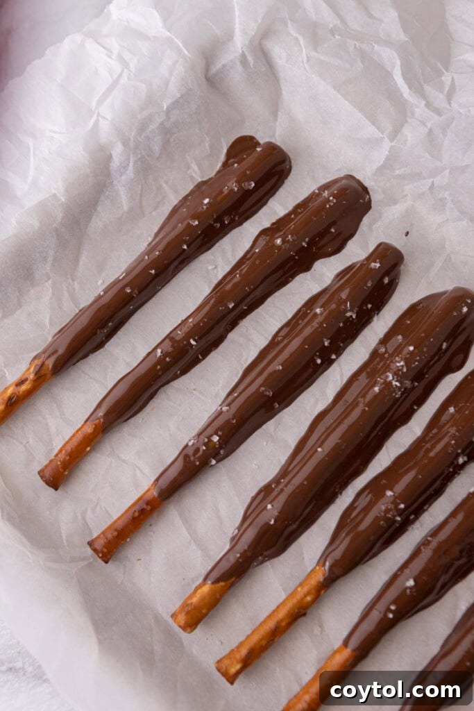 Chocolate-covered pretzel rods, with caramel beneath, resting on a sheet, some adorned with sprinkles, waiting to set.