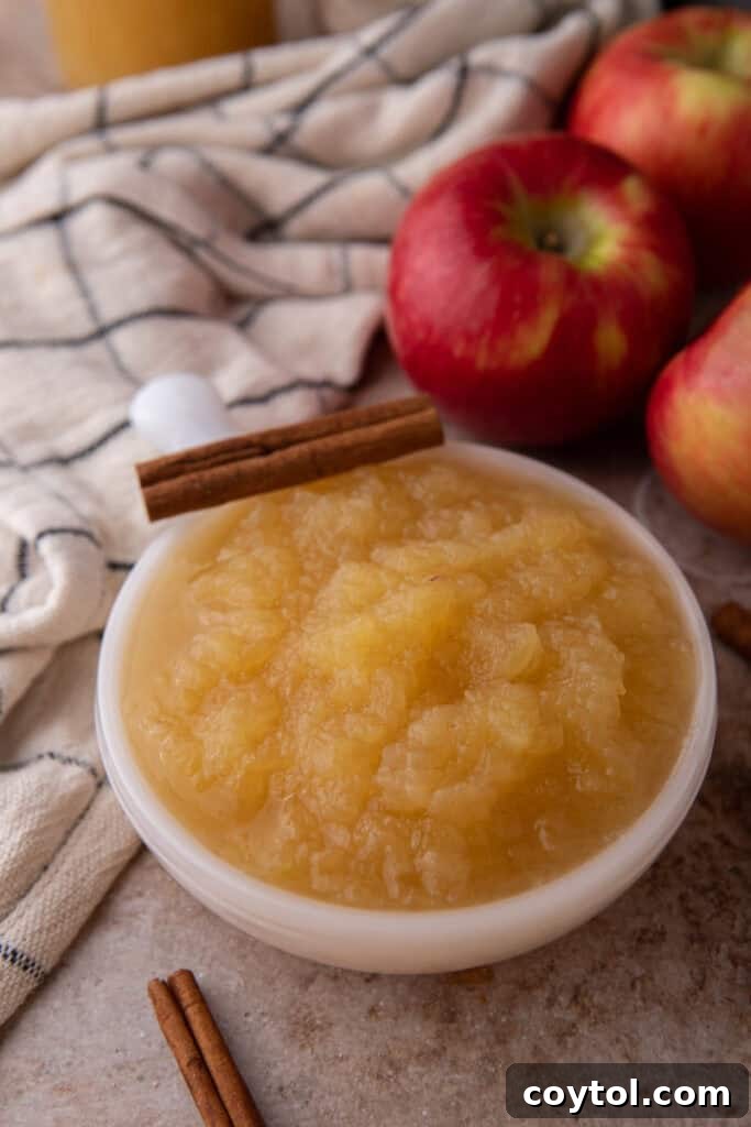 A clear glass bowl filled with Instant Pot Applesauce, smooth and golden, artfully placed on a wooden board amidst fresh apples, evoking a rustic fall setting.