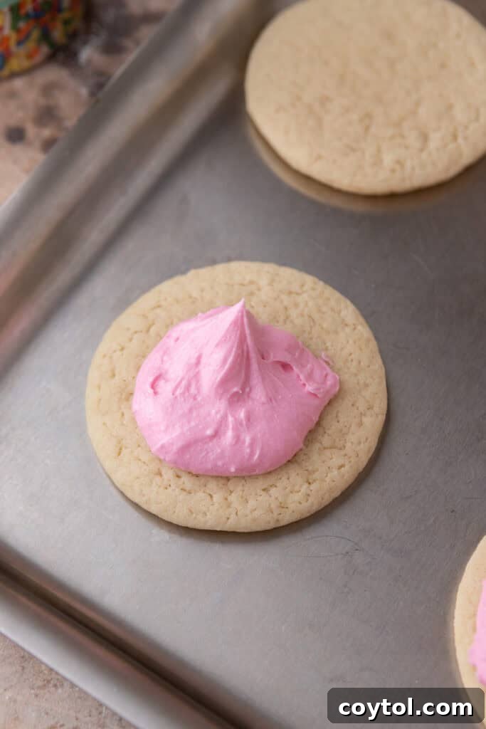 Pillowy Old-Fashioned Sour Cream Sugar Cookies 11 A baked sour cream sugar cookie topped with a spoonful of fluffy pink frosting before spreading.