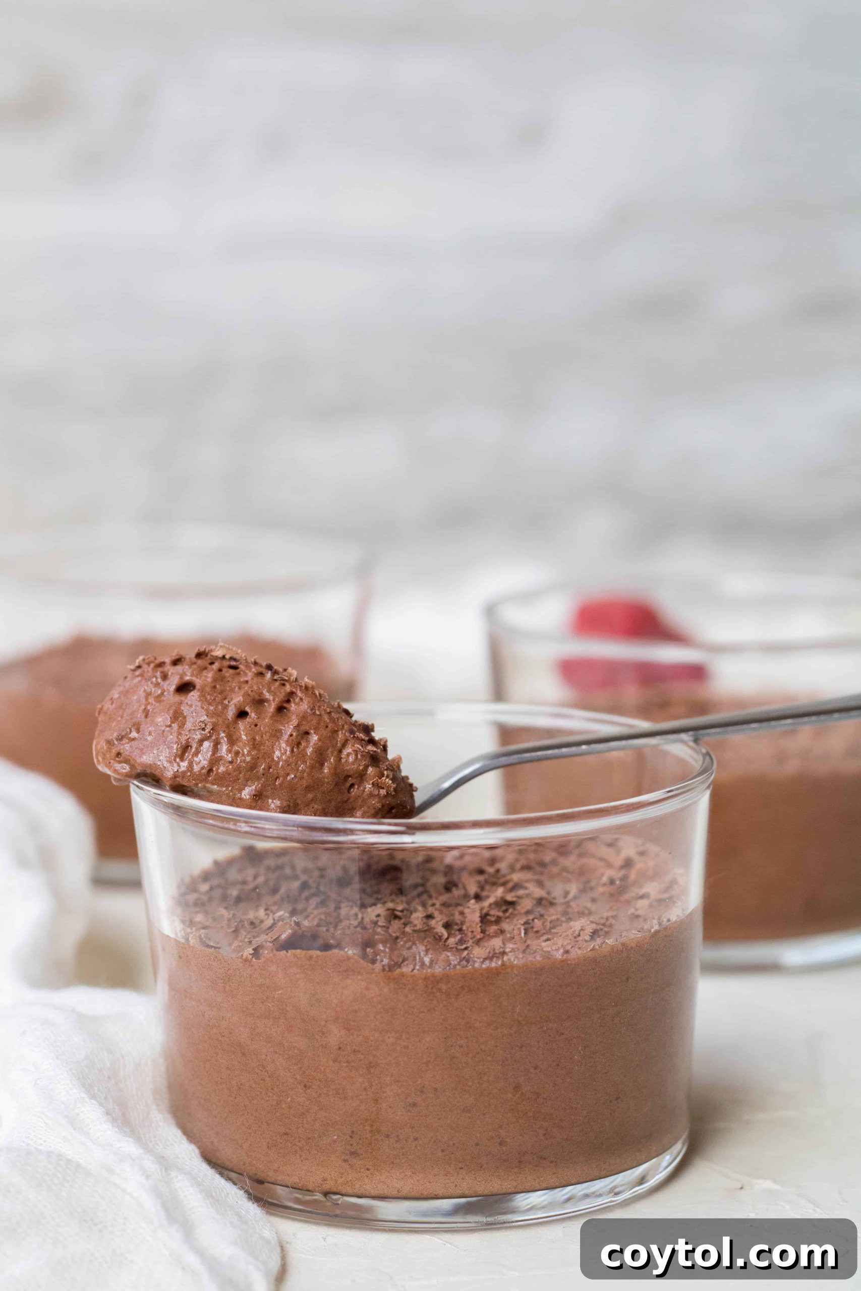 A delicate serving of vegan chocolate mousse, adorned with grated chocolate and vibrant fresh raspberries, in a small glass cup.