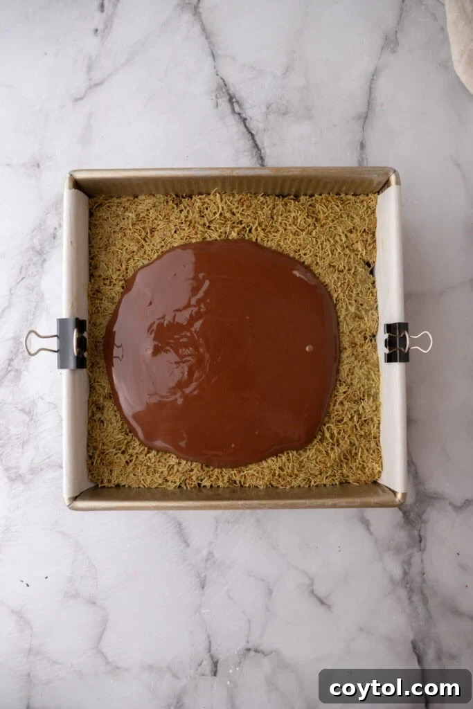 Glossy melted chocolate topping being spread over the pistachio kataifi layer.