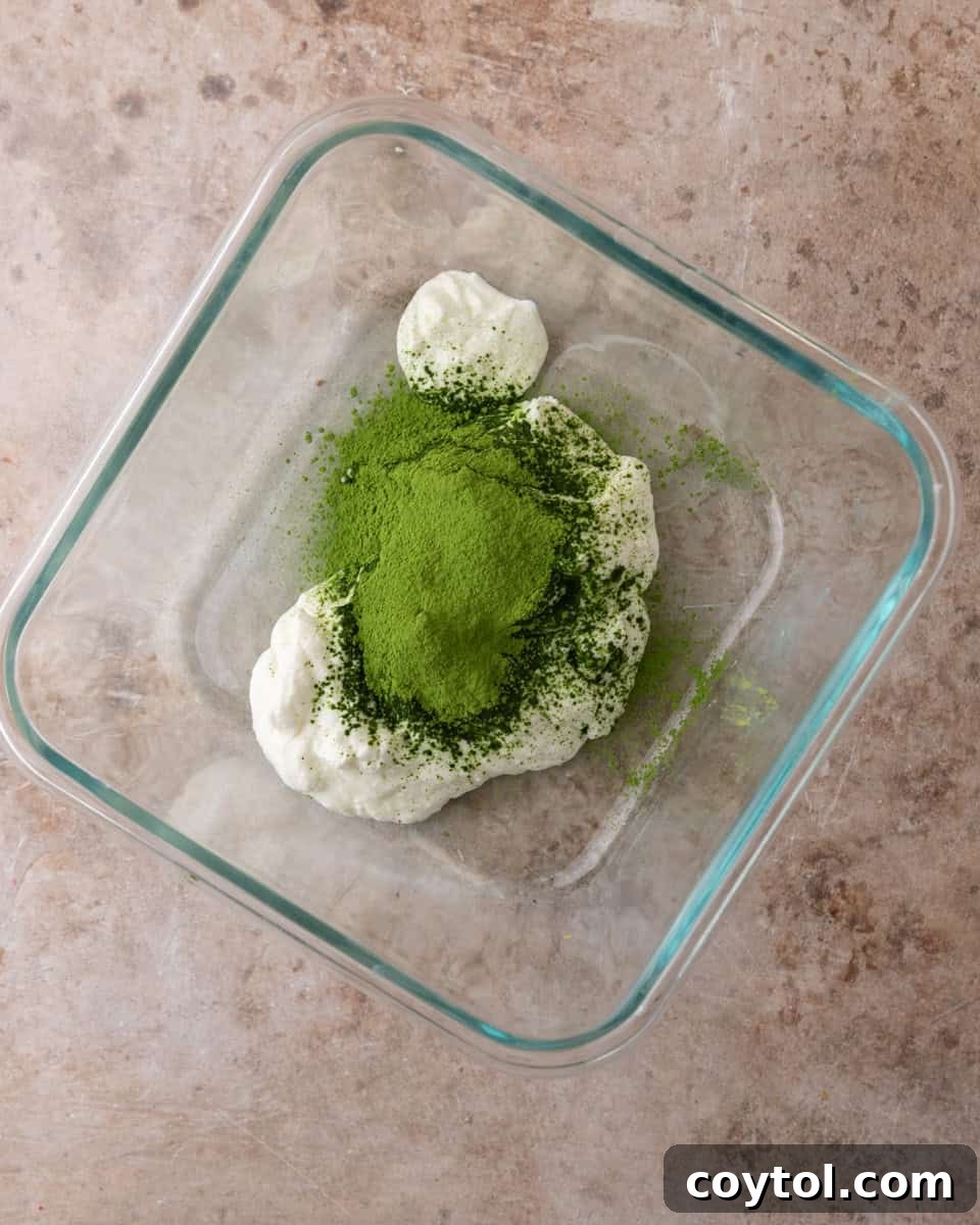 Matcha powder vigorously whisked into plain Greek yogurt in a bowl, creating a smooth green paste.