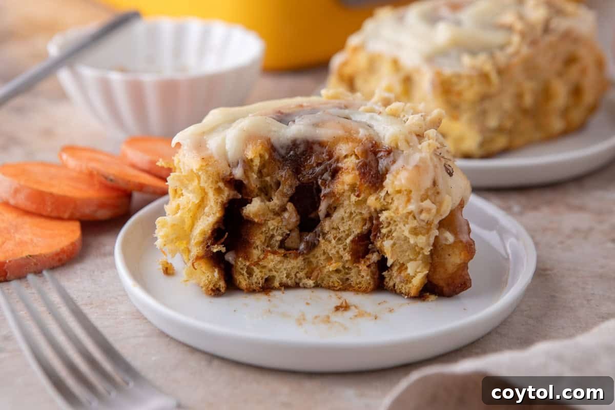 A carrot cake cinnamon roll with brown butter cream cheese frosting on a white plate with a bite taken out, showing the soft fluffy interior and spiced filling