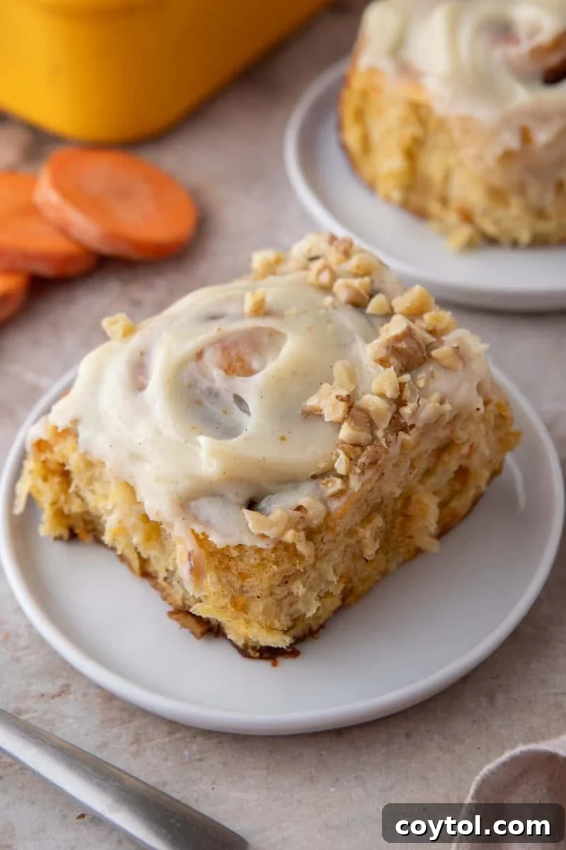 A frosted carrot cake cinnamon roll topped with chopped walnuts on a white plate with sliced carrots and a yellow baking pan in the background