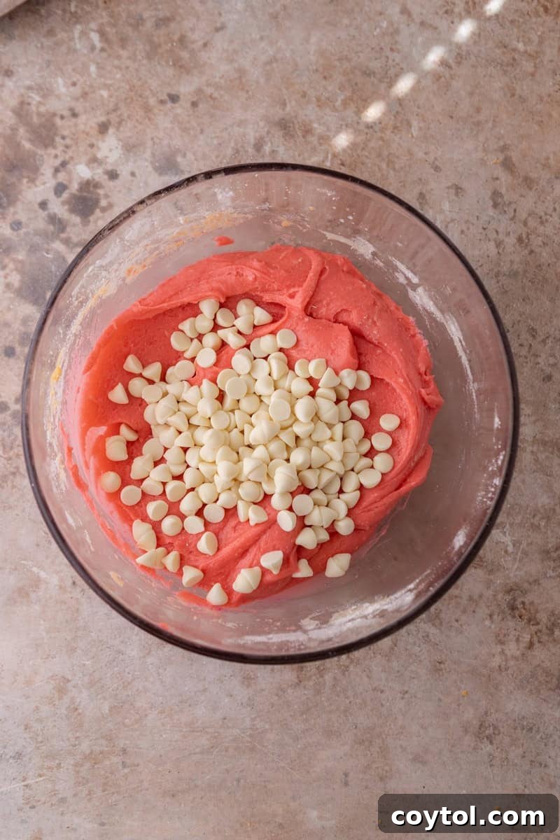 Bright pink strawberry cookie dough in a glass bowl with white chocolate chips on top ready to be folded in