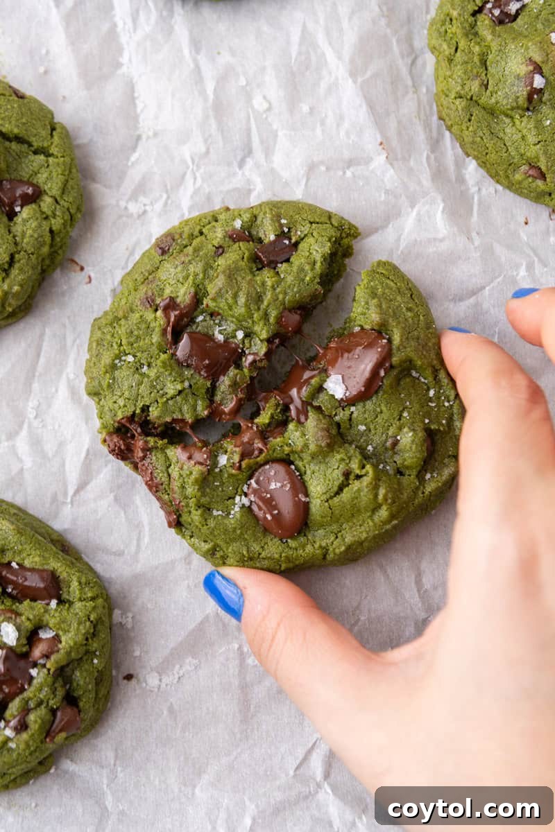 Hand breaking apart a thick chewy matcha cookie with melted chocolate and sea salt flakes on parchment paper