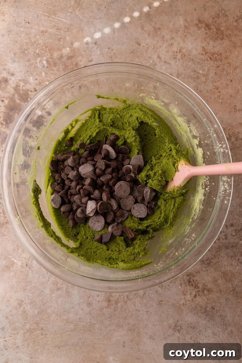 Bright green cookie dough with semi-sweet chocolate chips being folded in with a pink spatula
