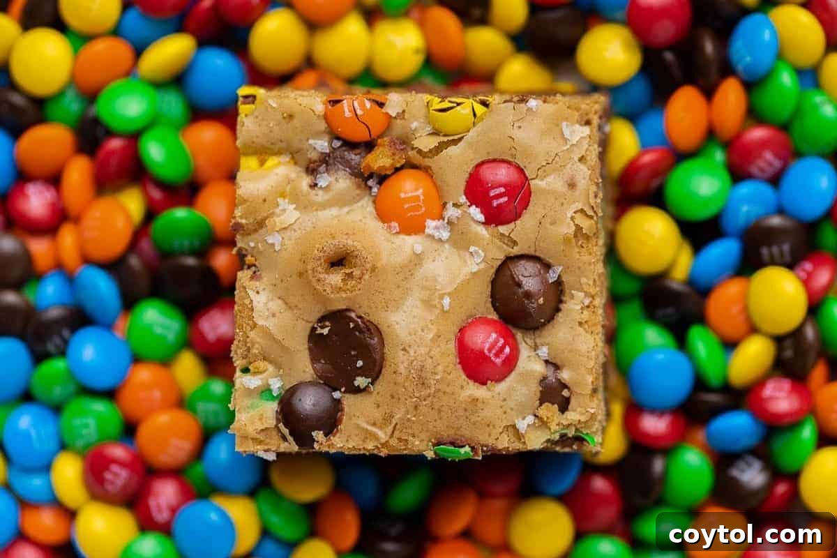 A single M&M chocolate chip blondie bar, beautifully surrounded by an assortment of colorful M&M candies, clearly showing its chewy texture and the delightful baked-in candy pieces, perfect for parties.