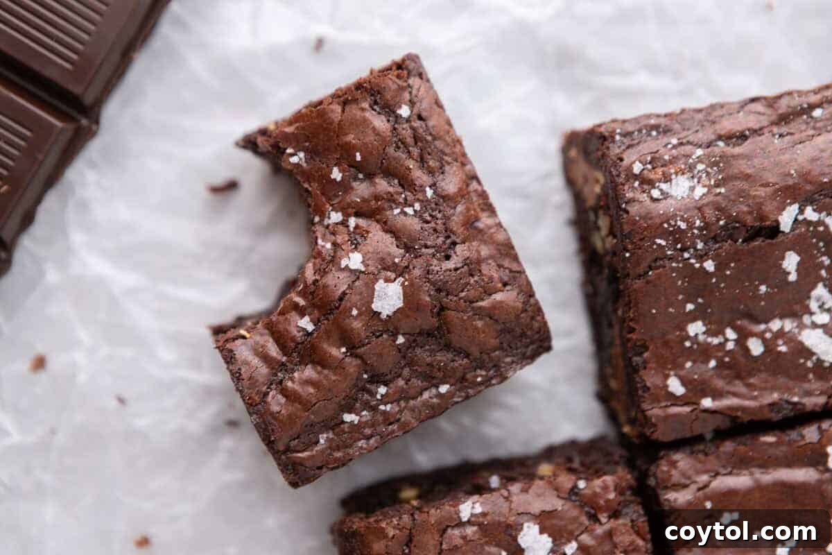 Overhead view of fudgy homemade brownies with crinkly tops and flaky sea salt on parchment paper next to dark chocolate squares, showcasing their rich, dense texture and perfect bake.