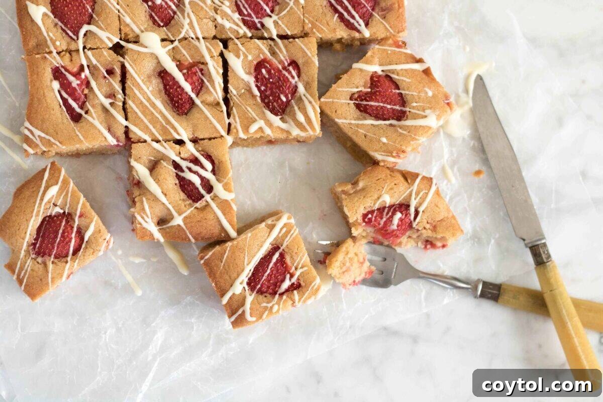 Exquisite brown butter strawberry blondies, delicately drizzled with white chocolate and presented on parchment paper, featuring fresh strawberry halves beautifully baked into each square, a gorgeous summer treat.