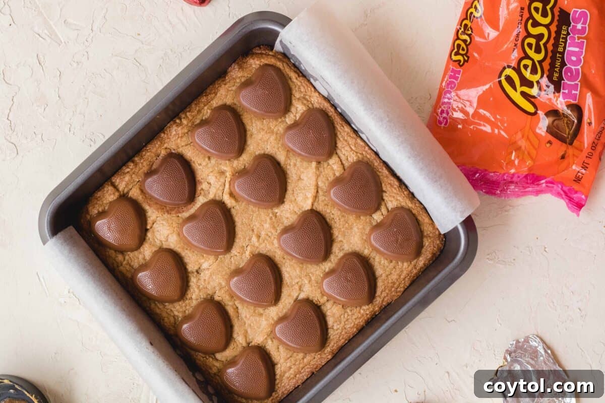 Rich peanut butter blondies baking in a pan, generously topped with iconic Reese's peanut butter hearts, with the candy bag alongside, ideal for Valentine's Day.
