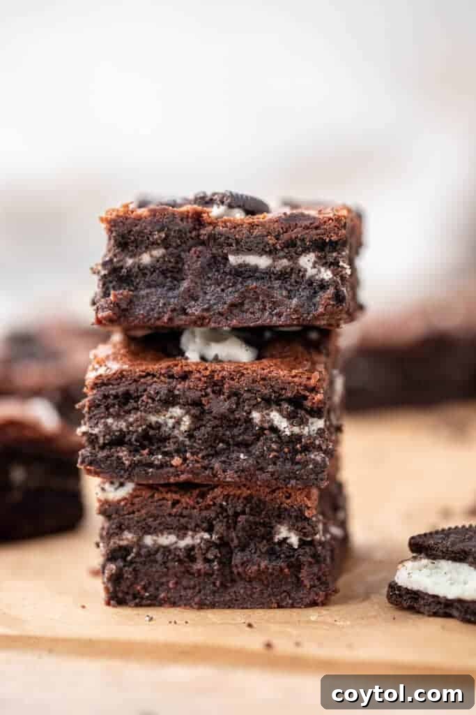 Stack of three fudgy Oreo brownies, clearly showing layers of iconic Oreo cookies baked into the dense chocolate interior, with extra Oreo pieces scattered on top, a cookies-and-cream lover's dream.