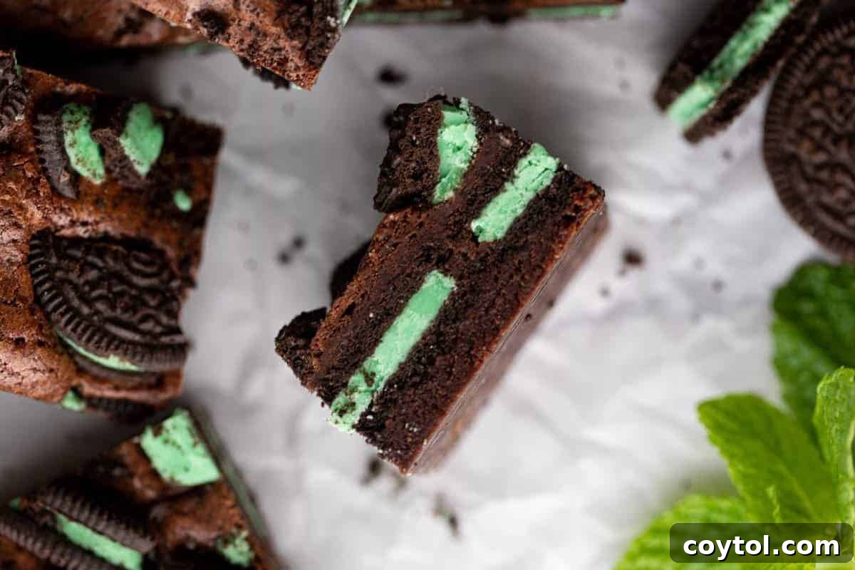 A tantalizing mint Oreo brownie, perfectly tilted to reveal its cross-section, displaying vibrant layers of bright green mint Oreo filling baked into the fudgy chocolate brownie, artfully surrounded by fresh mint leaves and mint Oreo cookies, perfect for St. Patrick's Day.