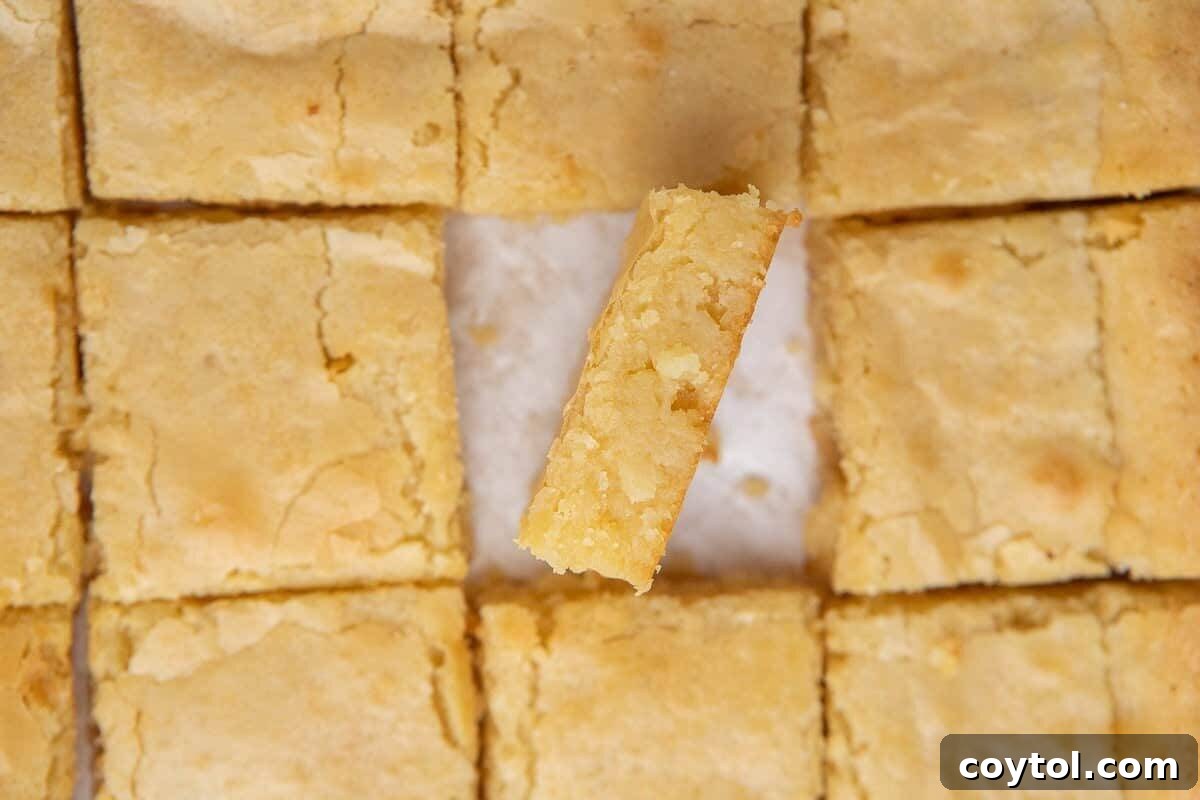 White chocolate brownies, neatly sliced into squares, with one piece gently tilted to reveal its dense, fudgy golden interior and a lovely crinkly top, offering a non-traditional brownie experience.