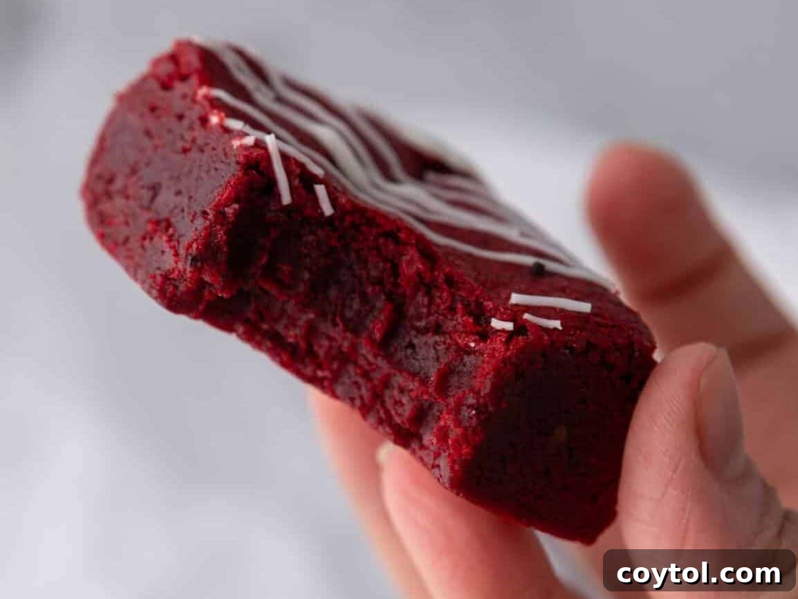 A hand holds a fudgy red velvet brownie with a bite taken out, revealing its dense, vibrant red interior and a delicate white chocolate drizzle on top, perfect for Christmas or Valentine's.