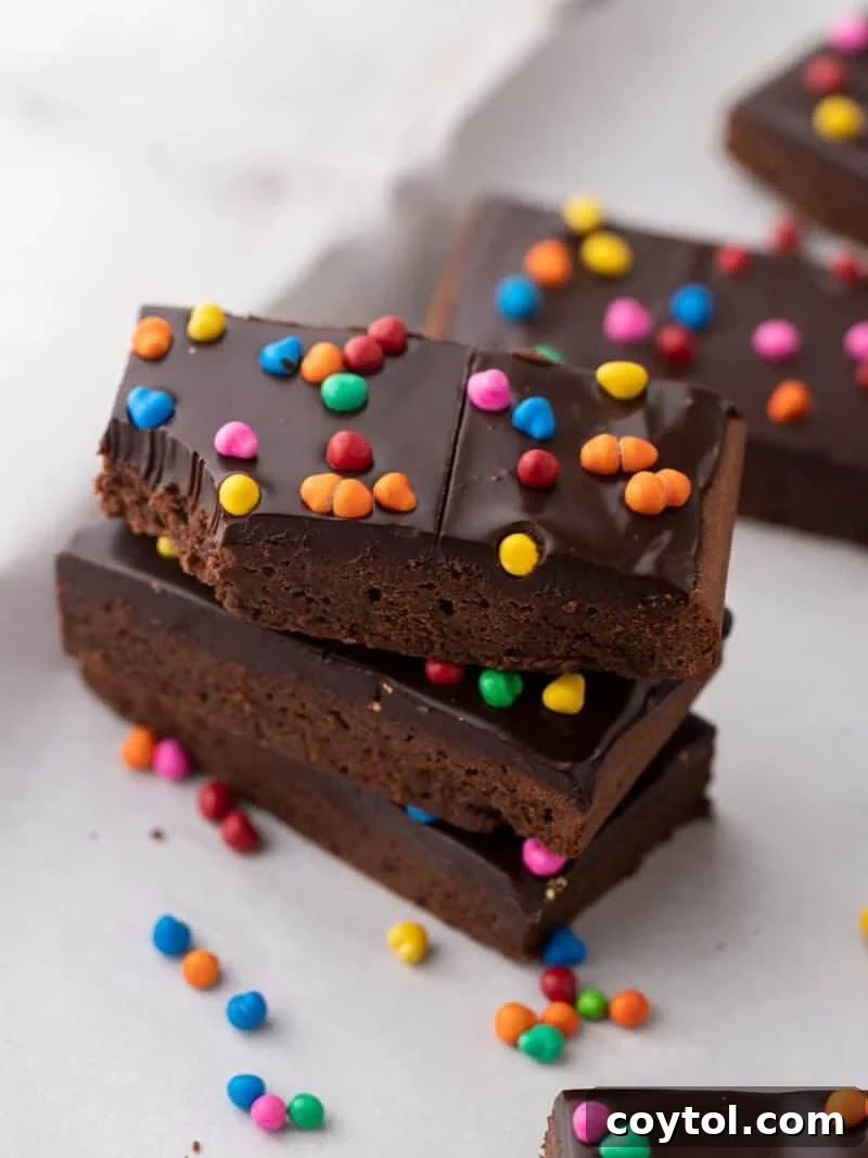 A stack of three homemade cosmic brownies, featuring a thick chocolate ganache and vibrant colorful rainbow candy chips on top, with extra candies scattered around, pure childhood nostalgia made from scratch.