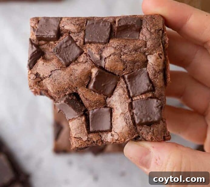 A hand holds a Starbucks copycat double chocolate brownie, generously loaded with chocolate chunks on top, with a cup of coffee and more brownies blurred in the background, better than the original.