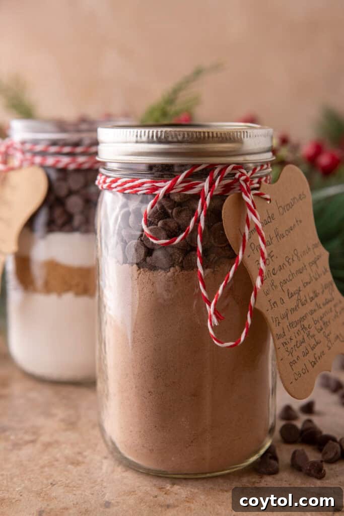 Two charming mason jars, elegantly filled with layered homemade brownie mix dry ingredients, tied with festive red and white baker's twine and adorned with handwritten recipe tags, against a backdrop of holiday greenery, ideal for gifting.