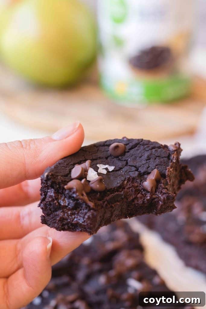 A hand holds a vegan black bean brownie with chocolate chips and flaky sea salt on top, showing the dense fudgy interior, with an avocado in the background, unbelievably fudgy despite black beans.