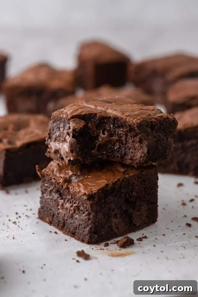 Two fudgy brown butter brownies stacked on parchment paper, with visible melted chocolate chunks inside and more brownies blurred in the background, highlighting their rich, nutty depth.