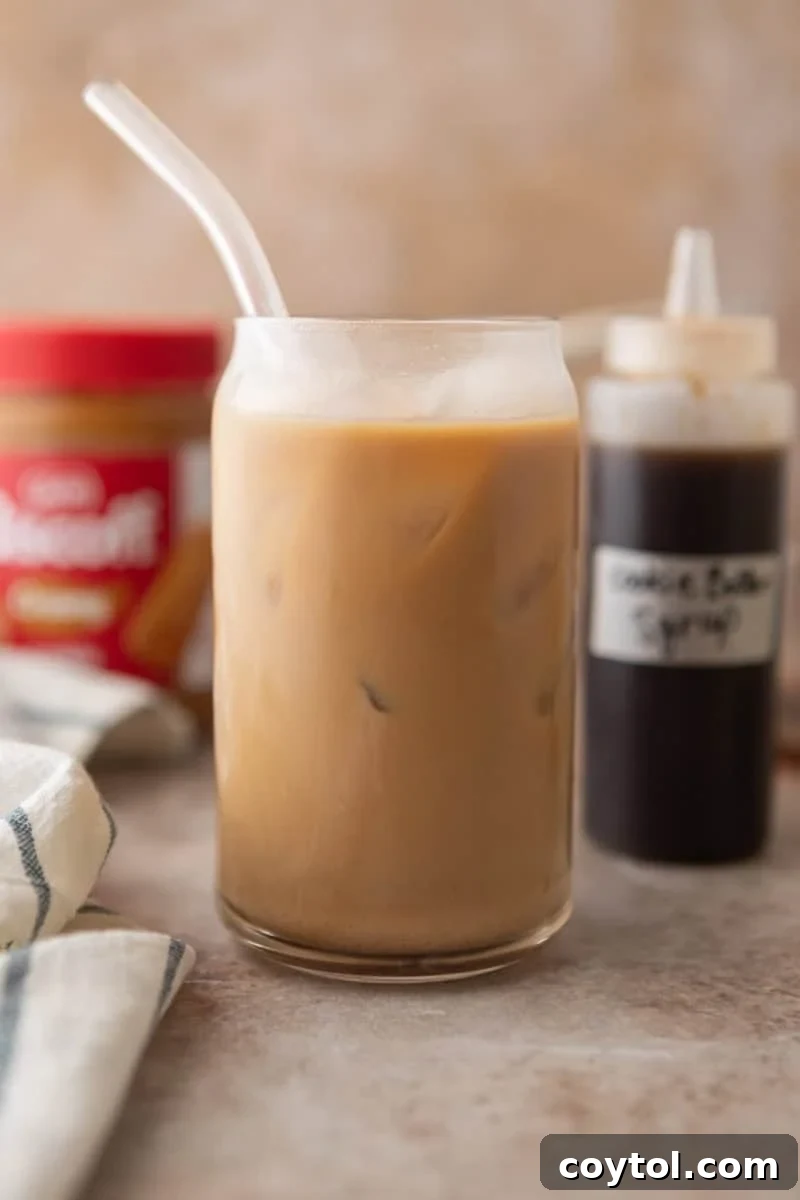 A cookie butter iced latte made with homemade Biscoff syrup in a glass with a straw, with the syrup bottle and Biscoff jar in the background.