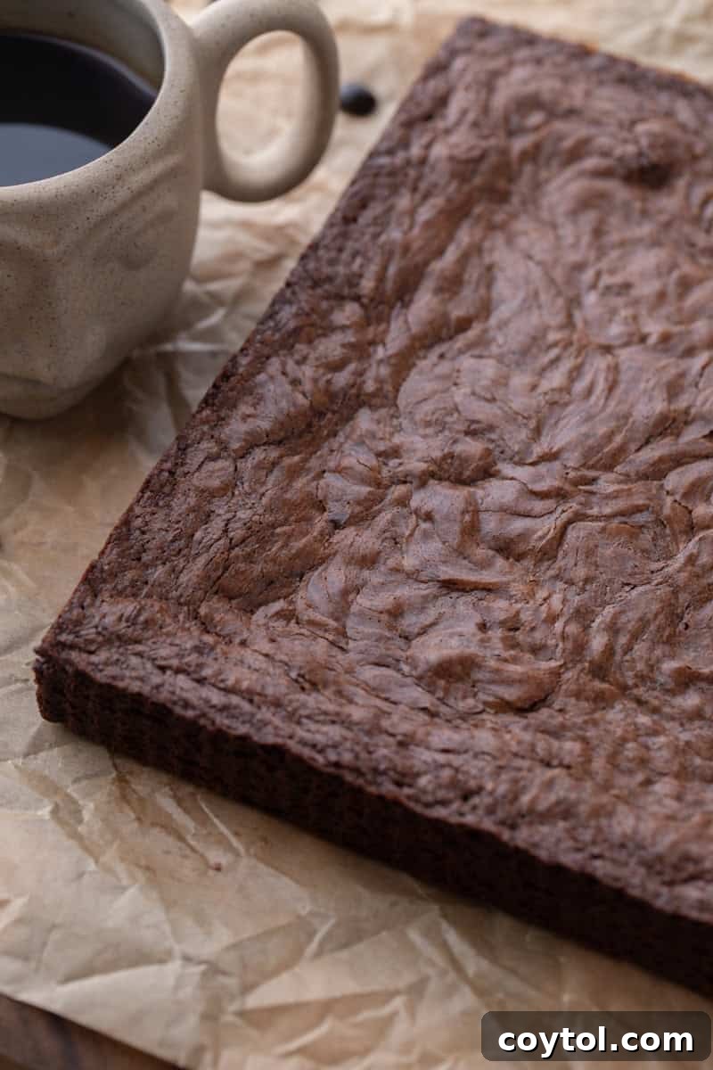 Whole slab of baked brownies with crinkly top on parchment next to a cup of coffee