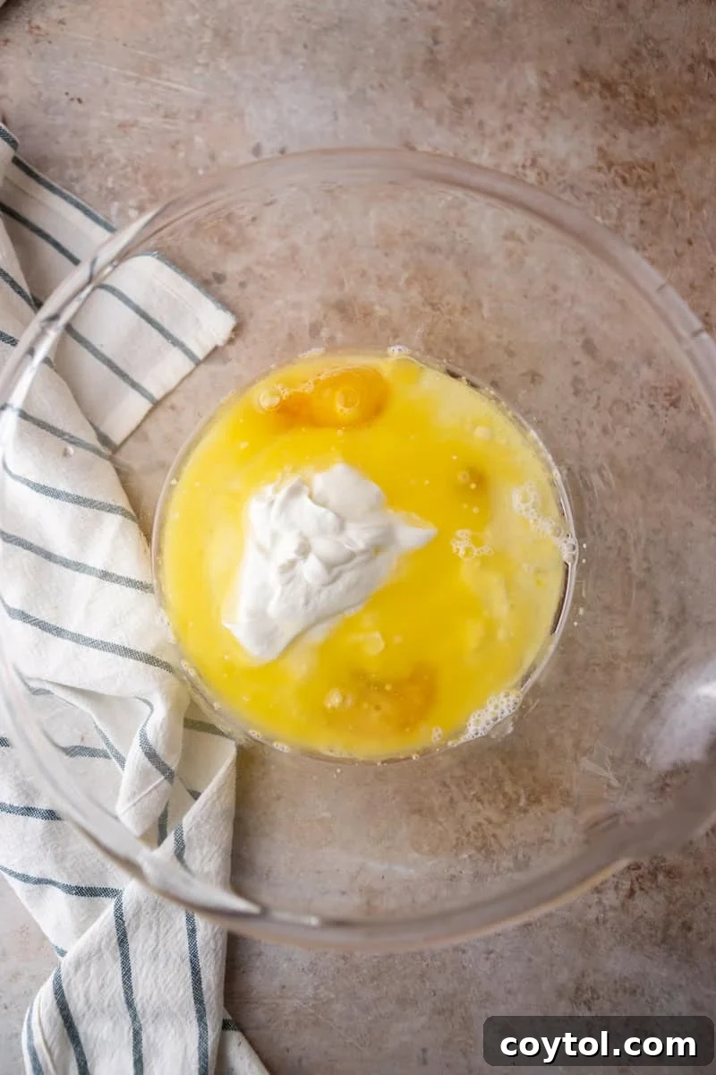 Eggs, melted butter, sour cream, and vegetable oil combined and whisked in a clear glass mixing bowl, showcasing the liquid components