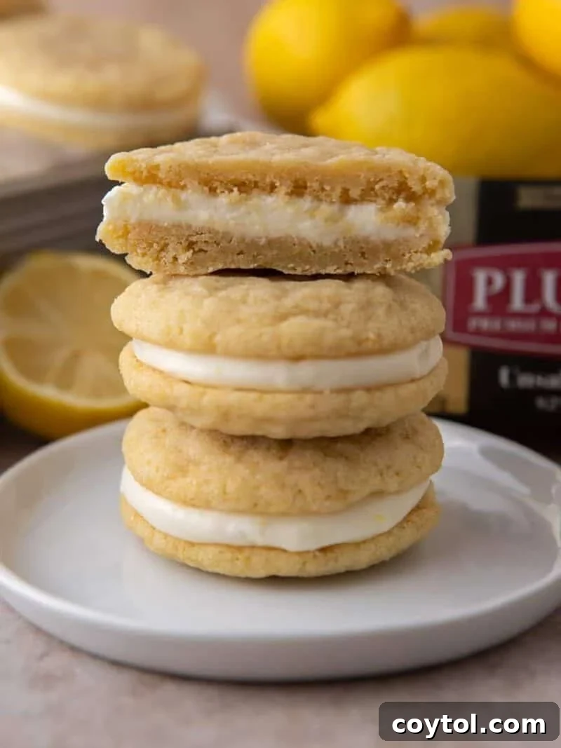 Stack of lemon sandwich cookies with cream cheese filling, top cookie split to show the inside, with lemons and Plugrà butter in the background.