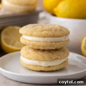 Two lemon sandwich cookies stacked on a small white plate with fresh lemons in the background.