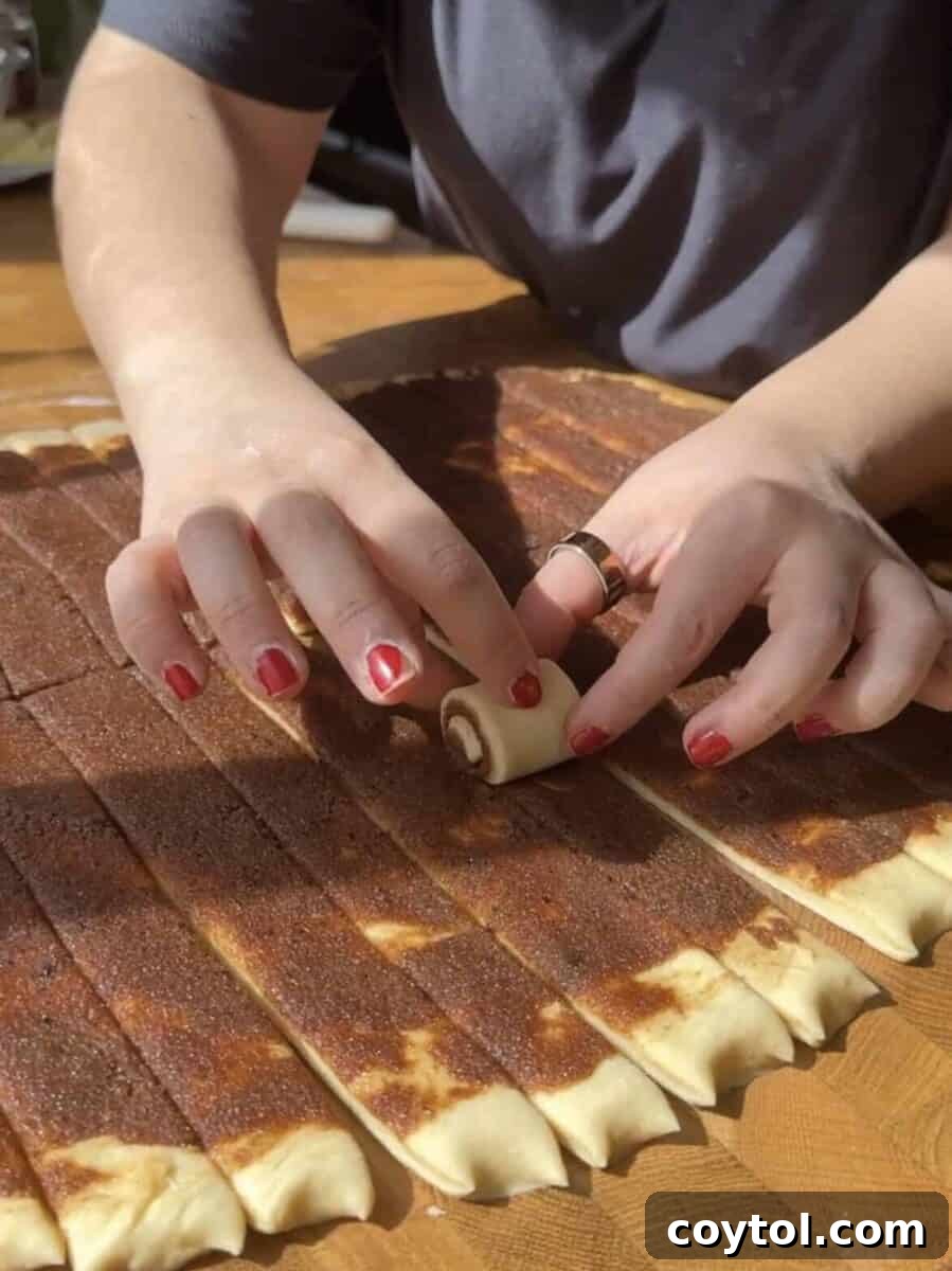 A hand is shown carefully rolling one of the small strips of dough into a tight mini cinnamon roll spiral.