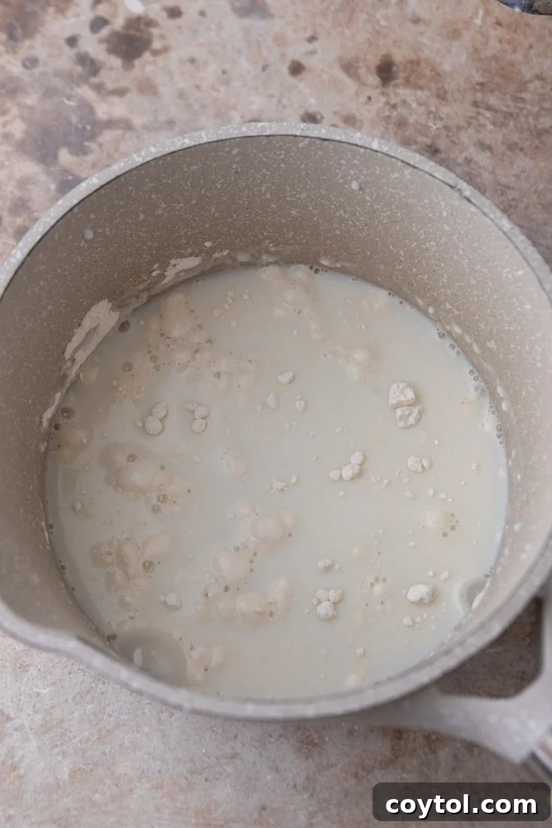 A whisk combines flour and milk in a small saucepan, creating the initial slurry for the tangzhong before cooking.