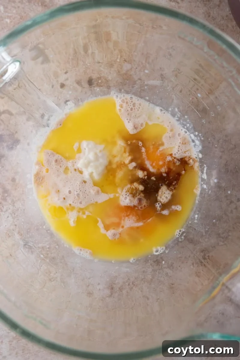 The wet ingredients for the cinnamon roll dough, including melted butter, eggs, cooled tangzhong, and the activated yeast mixture, are combined in a glass mixing bowl.