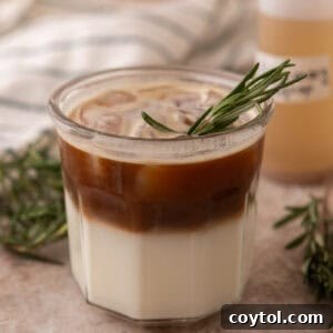 An iced rosemary latte layered in a glass, with a sprig of rosemary for garnish.