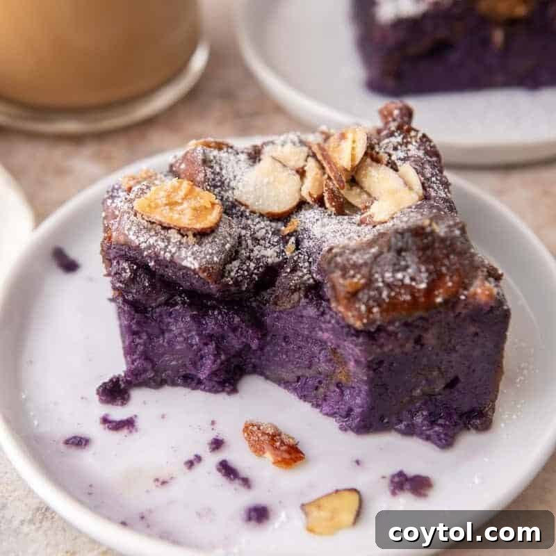 A square slice of vibrant purple ube French toast bake on a white plate, topped with powdered sugar and toasted almonds. A cup of coffee is visible in the background.