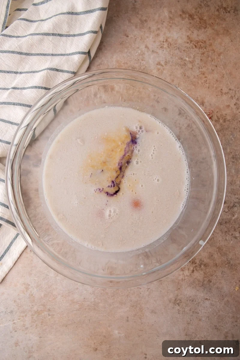 A glass bowl contains the ingredients for the ube custard before whisking: coconut milk, eggs, brown sugar, and purple ube extract.