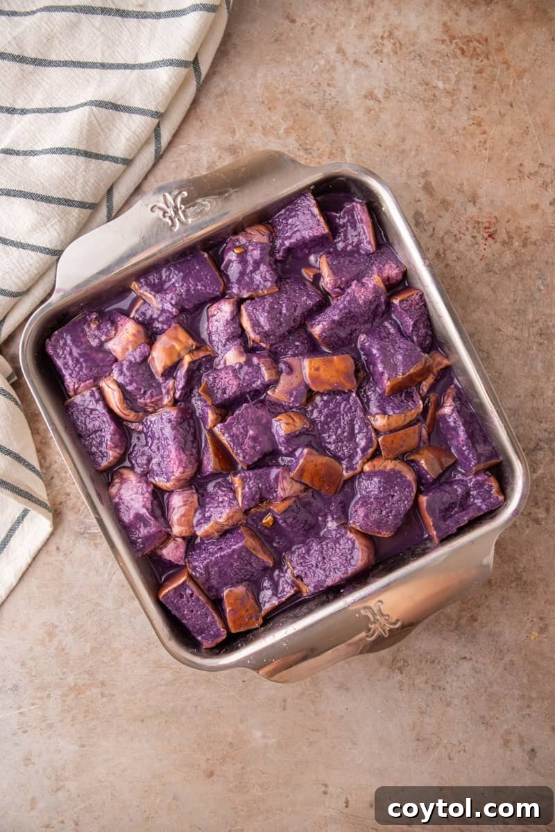Cubed Hawaiian rolls are soaking in the purple ube custard in a square metal baking dish, ready for refrigeration.