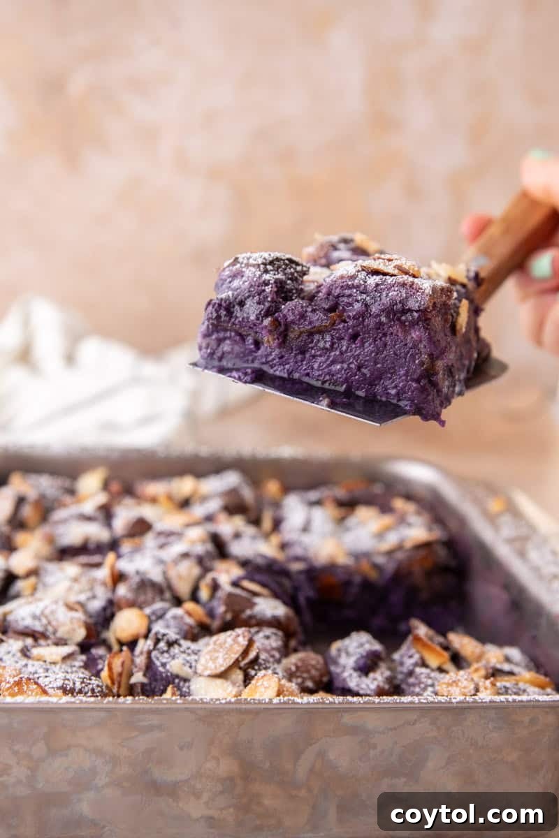 A spatula lifts a perfect square slice of the ube French toast bake from the pan, revealing the soft, custardy, and vibrant purple interior.