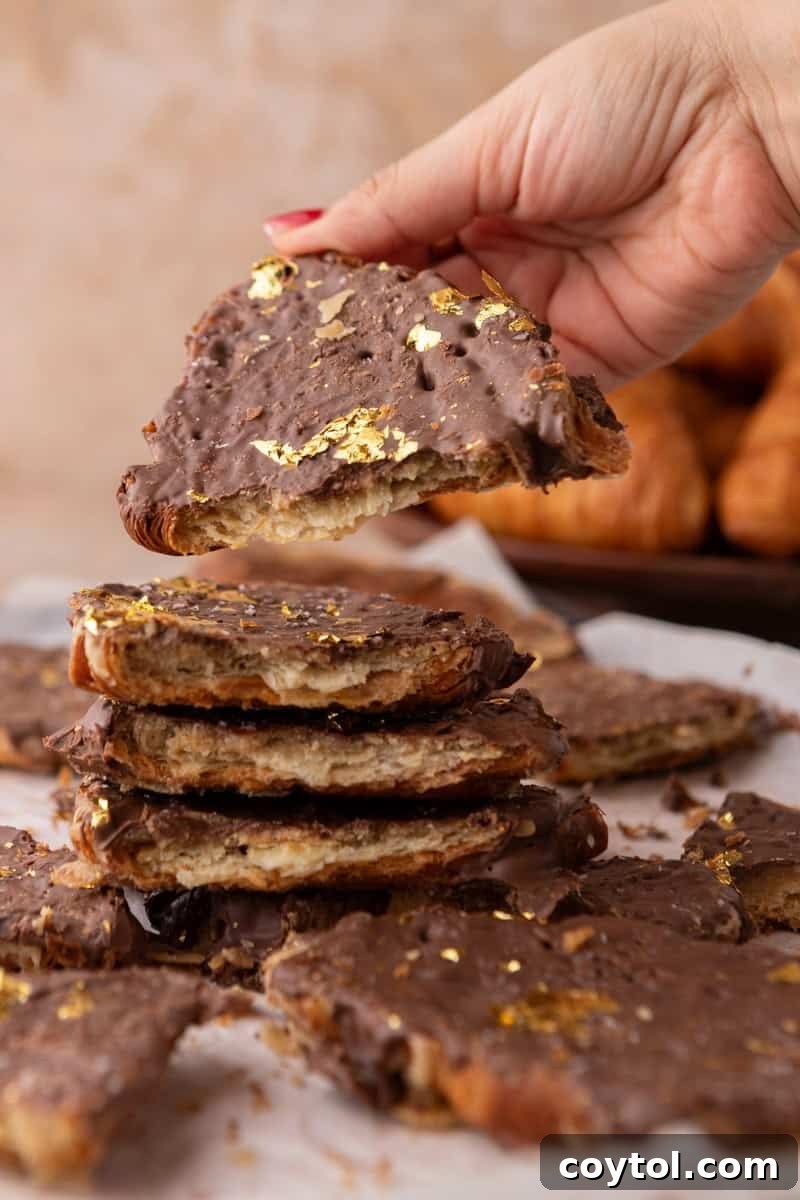 Hand holding a shard of croissant Christmas crack with chocolate and gold leaf above a stack of pieces