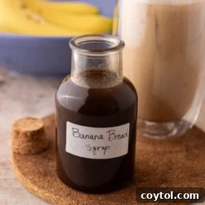 Banana bread syrup in a glass bottle with a handwritten label on a cork trivet next to an iced banana bread latte