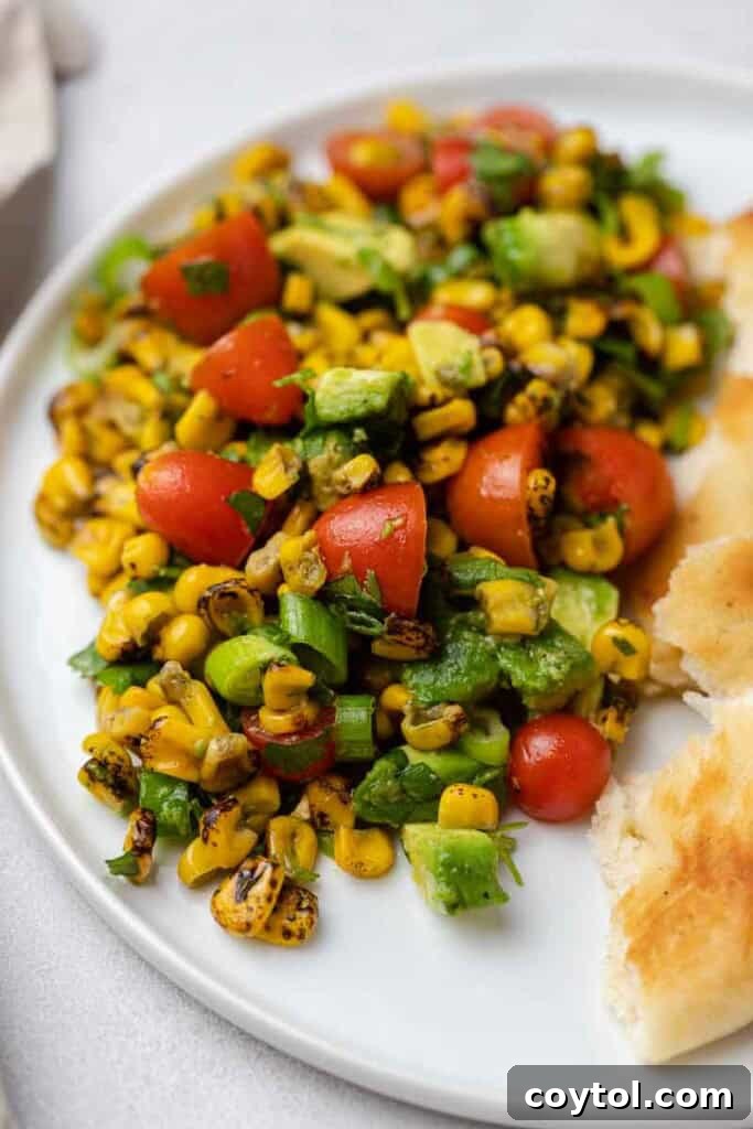 Roasted corn salad with avocado, cherry tomatoes, green onion, and herbs on a white plate with bread.