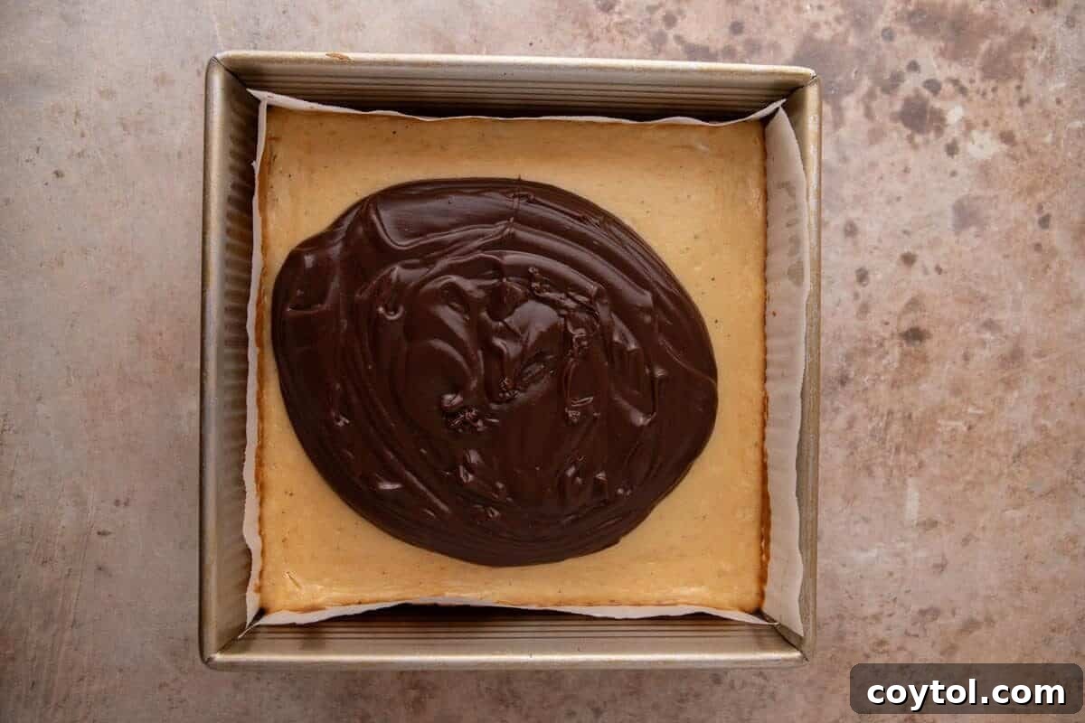 Chocolate ganache being spread over the baked marshmallow cheesecake layer for s'mores cheesecake bars