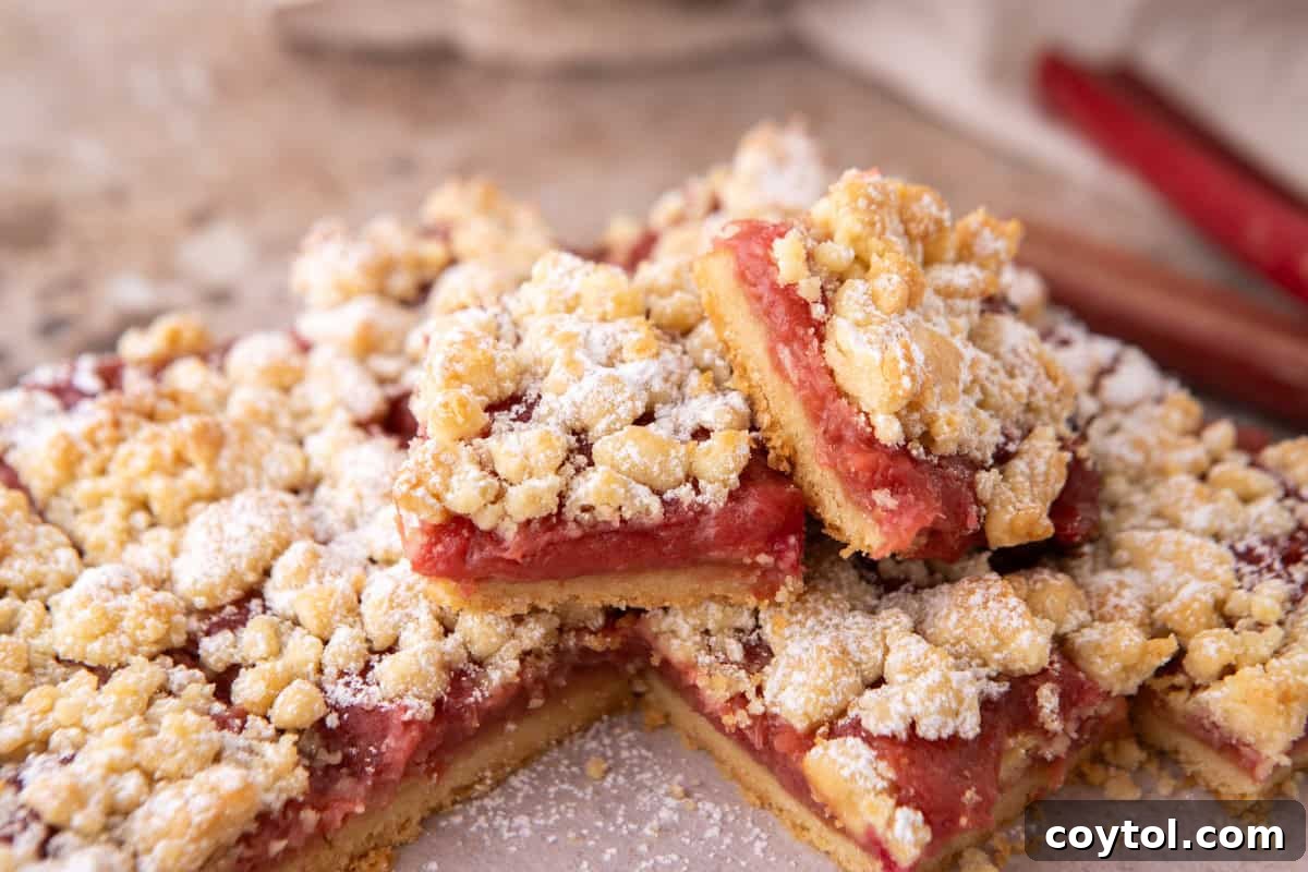 Rhubarb crumble bars stacked on a pink surface dusted with powdered sugar, with fresh rhubarb stalks in the background