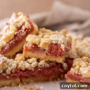 Stack of rhubarb crumble bars on parchment paper showing layers of golden shortbread base, pink jammy rhubarb filling, and crumble topping