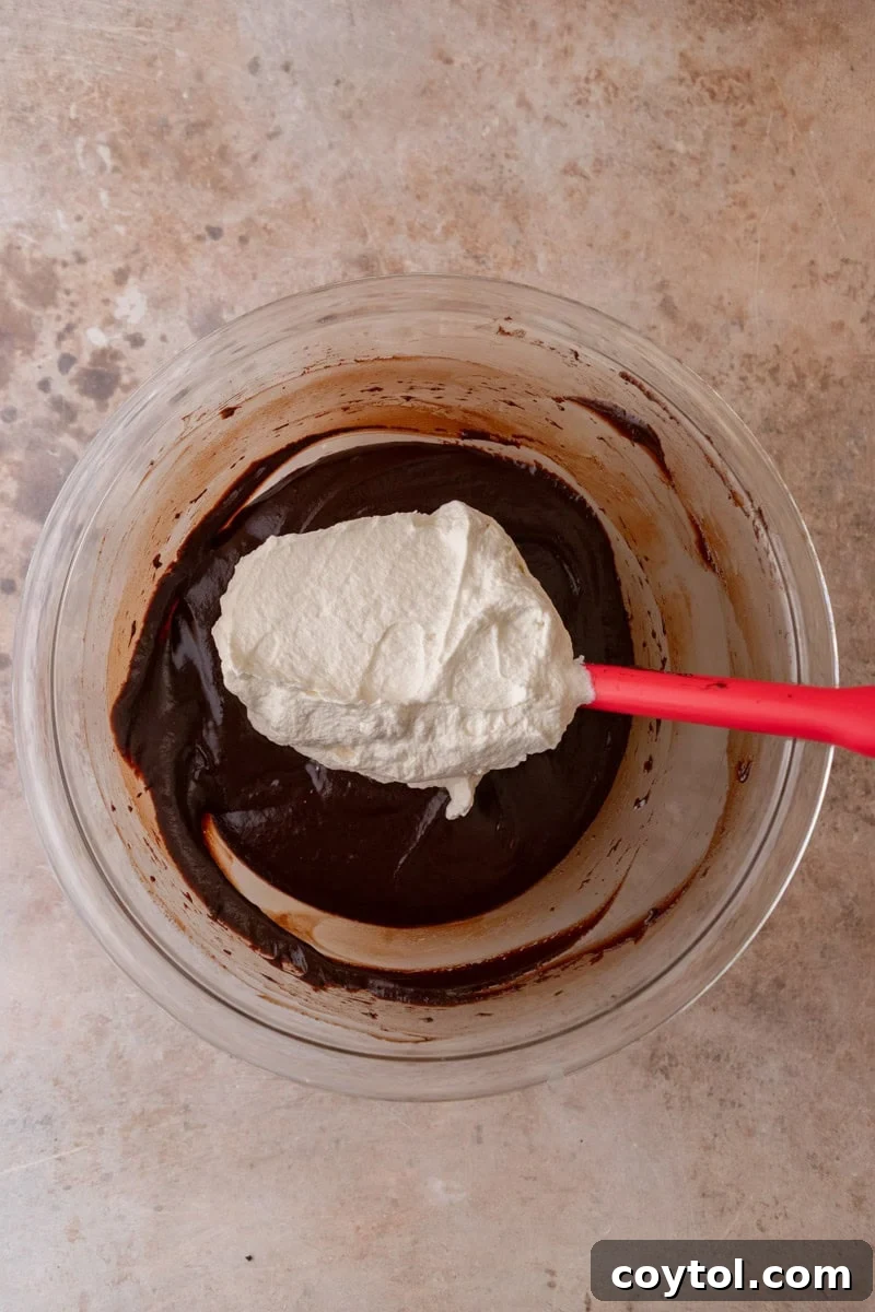 First addition of whipped cream being folded into the dark chocolate mixture with a red rubber spatula