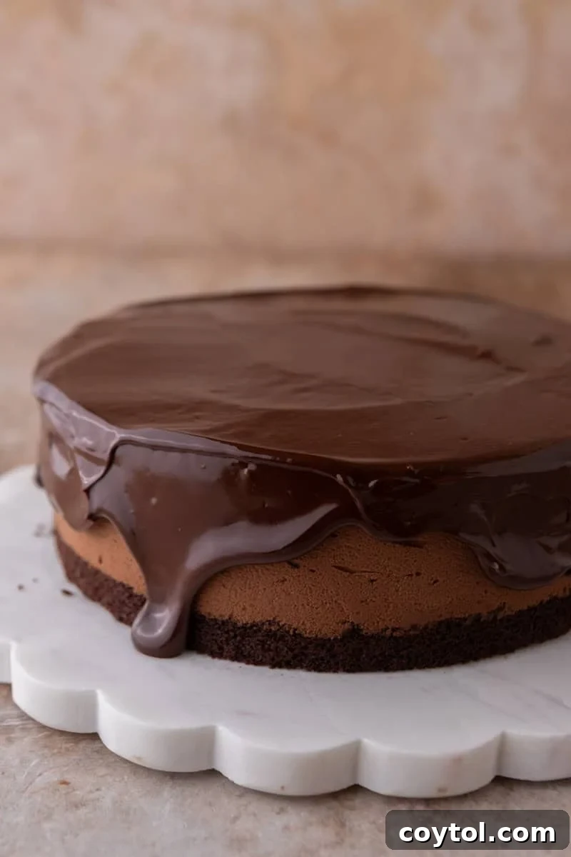 Chocolate ganache being poured over the chocolate mousse cake, dripping down the sides, on a white scalloped marble cake stand