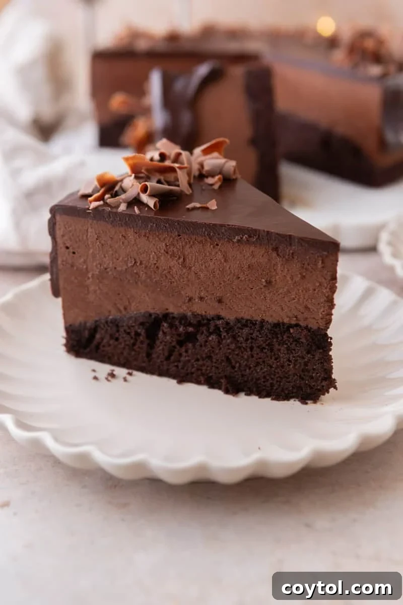A slice of chocolate mousse cake on a white scalloped plate showing three distinct layers of chocolate sponge cake, chocolate mousse, and chocolate ganache topped with chocolate curls