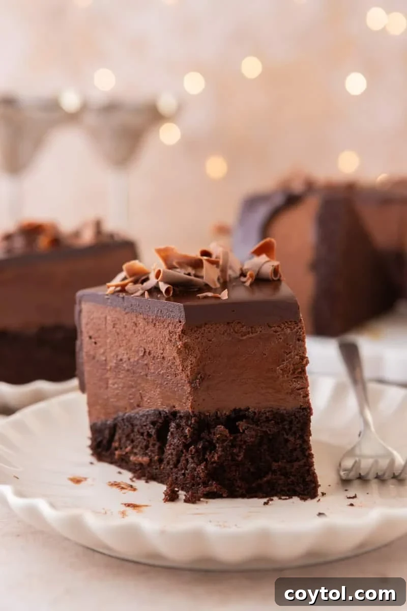 A slice of chocolate mousse cake on a plate with a fork, with the rest of the cake and soft bokeh lights in the background