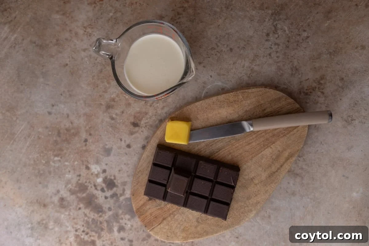 Ingredients for the chocolate ganache including heavy cream in a glass measuring cup, a dark chocolate bar, and butter on a wooden cutting board