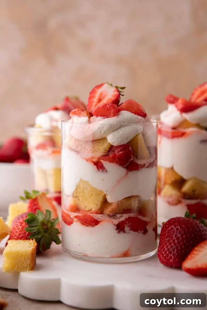 Three strawberry shortcake trifle cups layered with vanilla cake cubes, macerated strawberries, and whipped cream on a marble board.