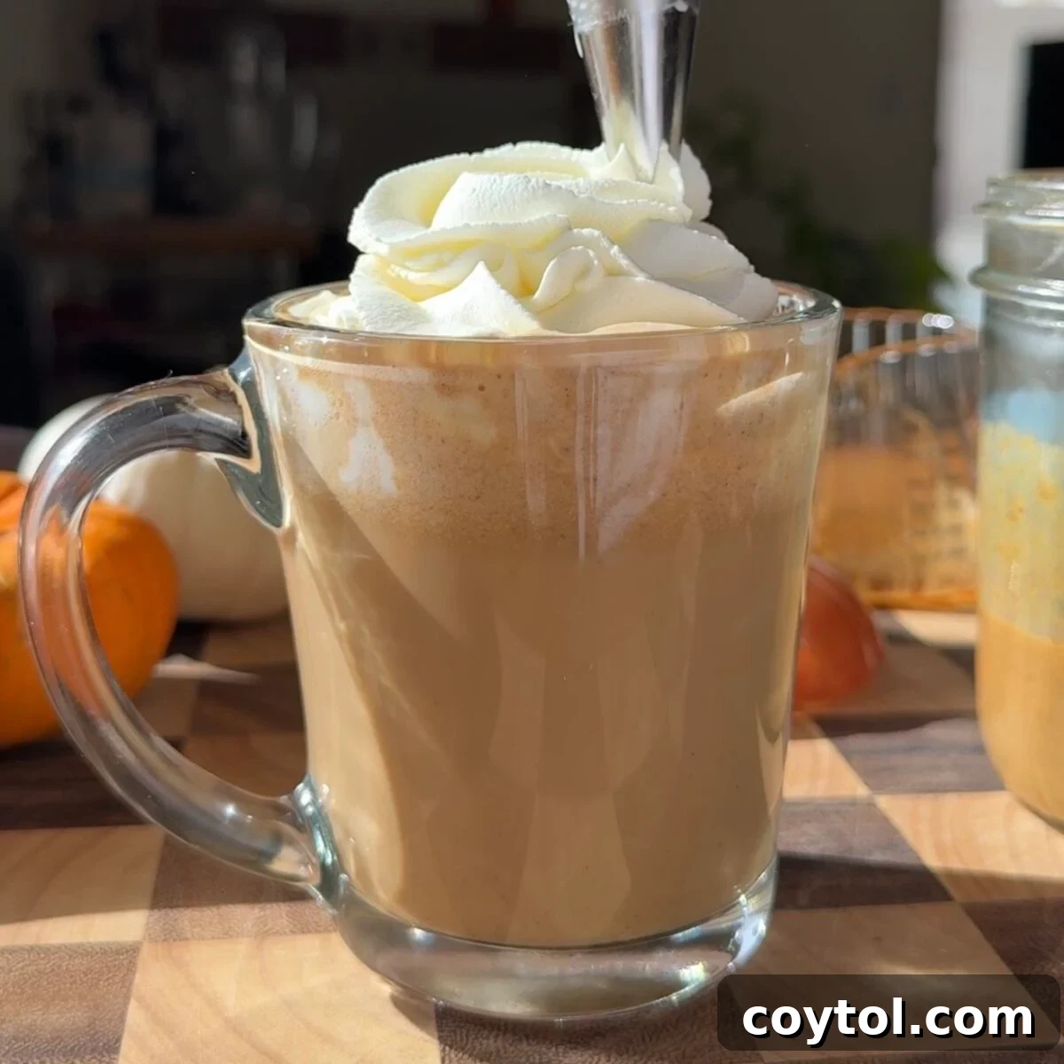 Whipped cream being piped on top of a pumpkin spice latte in a glass mug.
